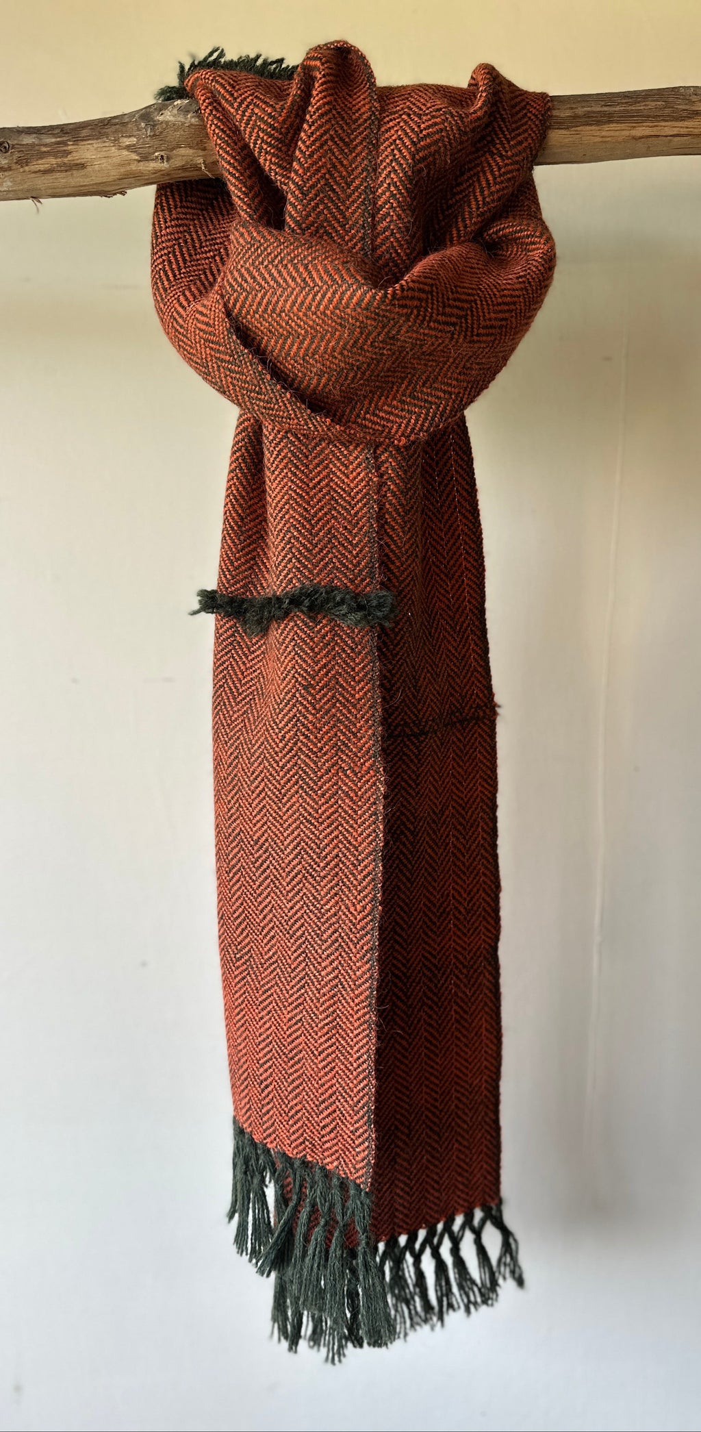 Burnt orange scarf with Dark green fringe hanging on a wooden stick against a white wall.