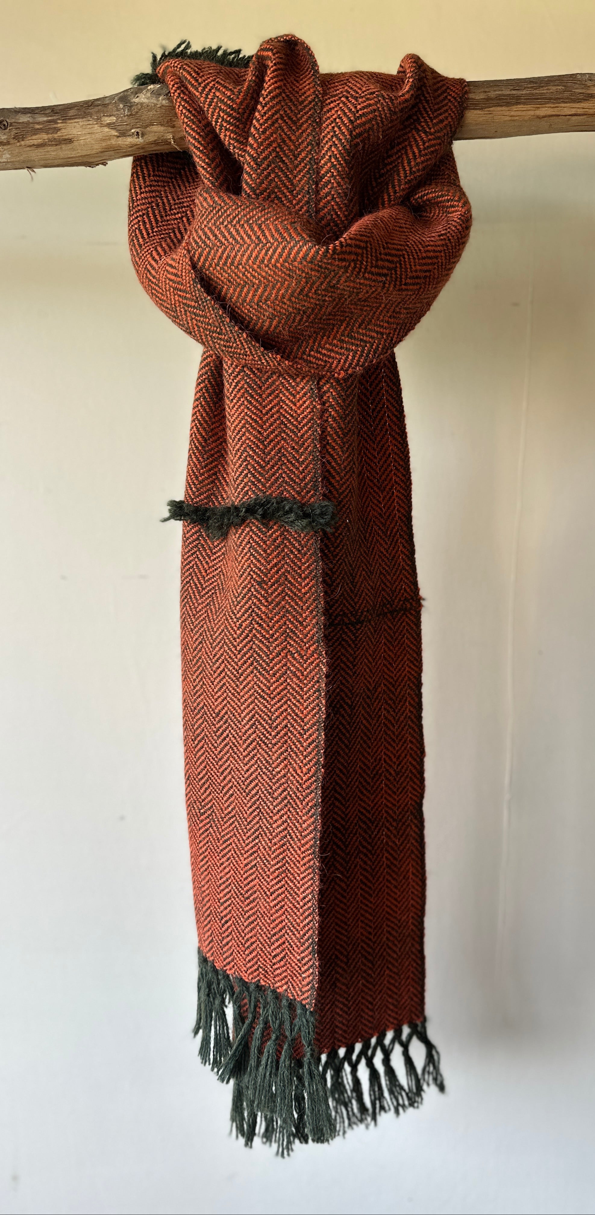 Burnt orange scarf with Dark green fringe hanging on a wooden stick against a white wall.