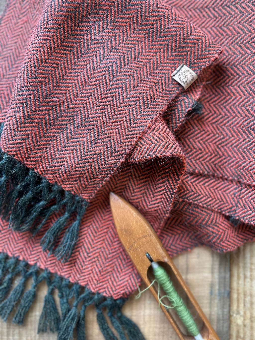 Burnt Orange herringbone scarf with Dark green fringes and a wooden spool of green thread on a textured surface.