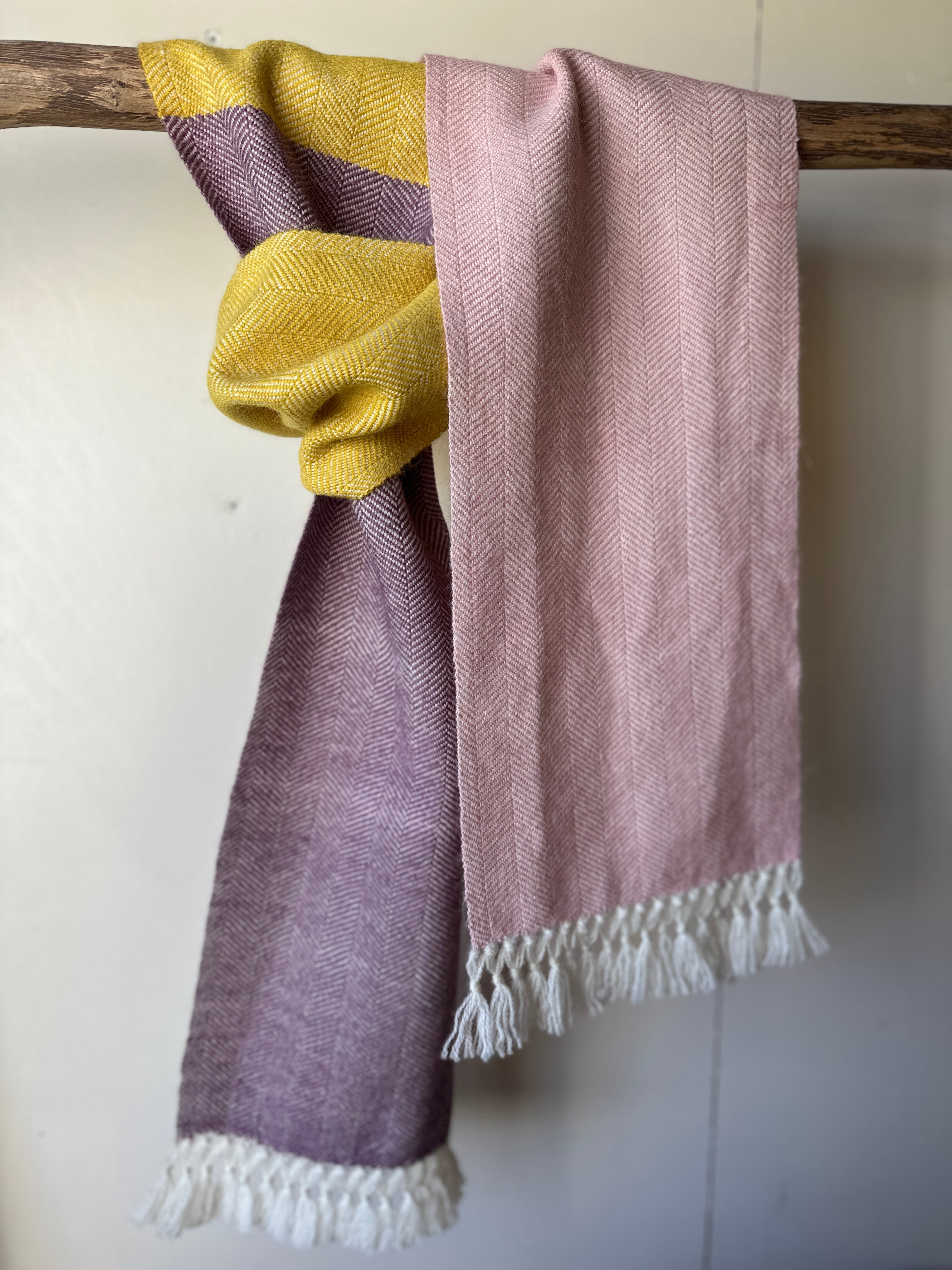 One herringbone scarf with three colours yellow, purple and pink with white fringe, draped over a wooden rod against a white background.