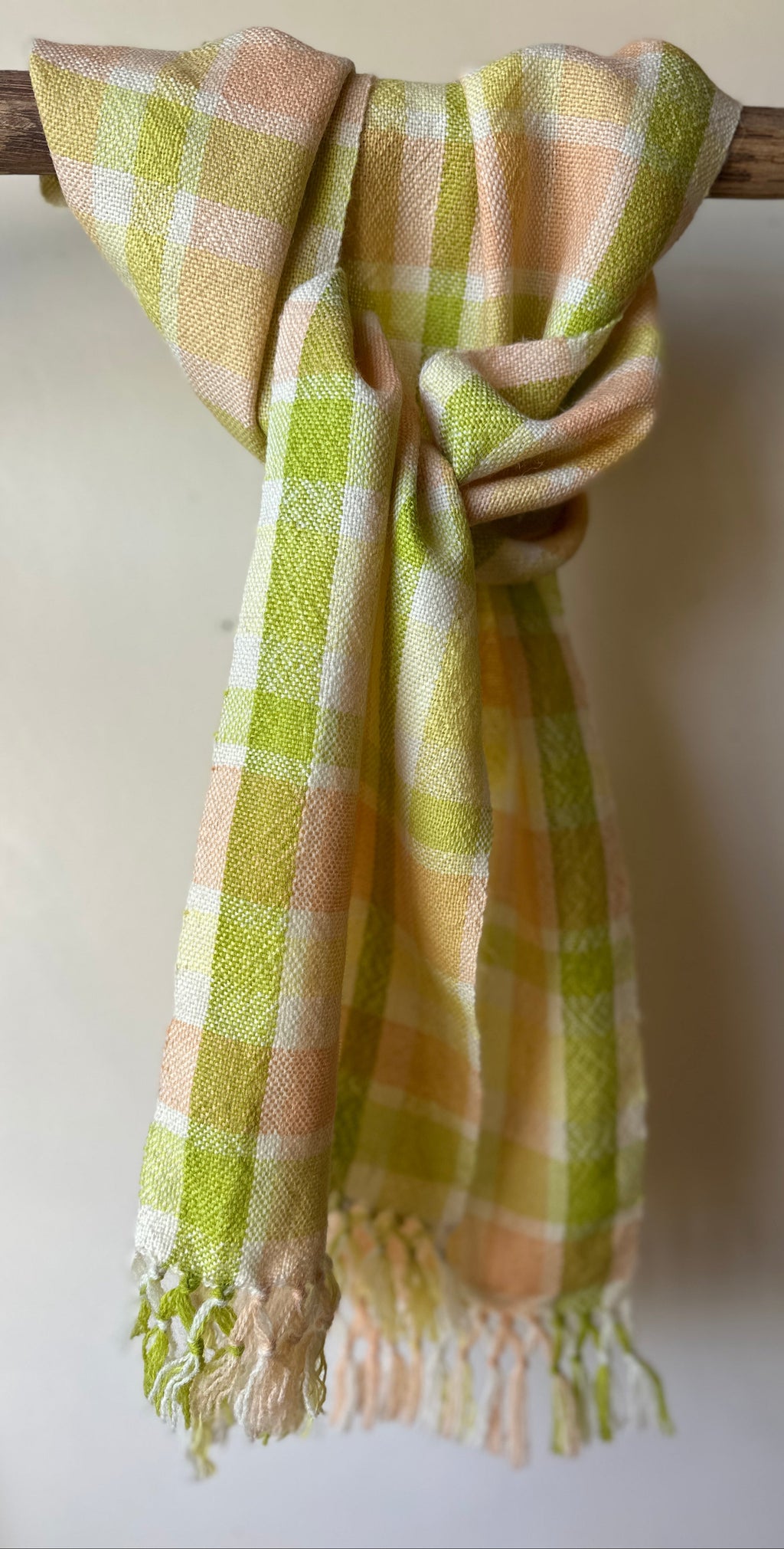 Plaid scarf with green and beige colors draped over a wooden stick against a white background